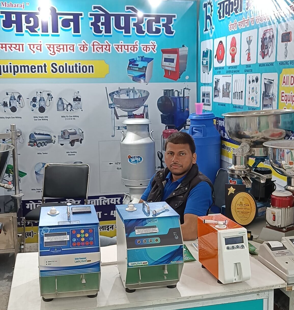 A man in a blue shirt sits behind a table displaying milk analyzers at a "Rakesh Cream Machine Separator" booth, with various dairy equipment shown on posters in the background.