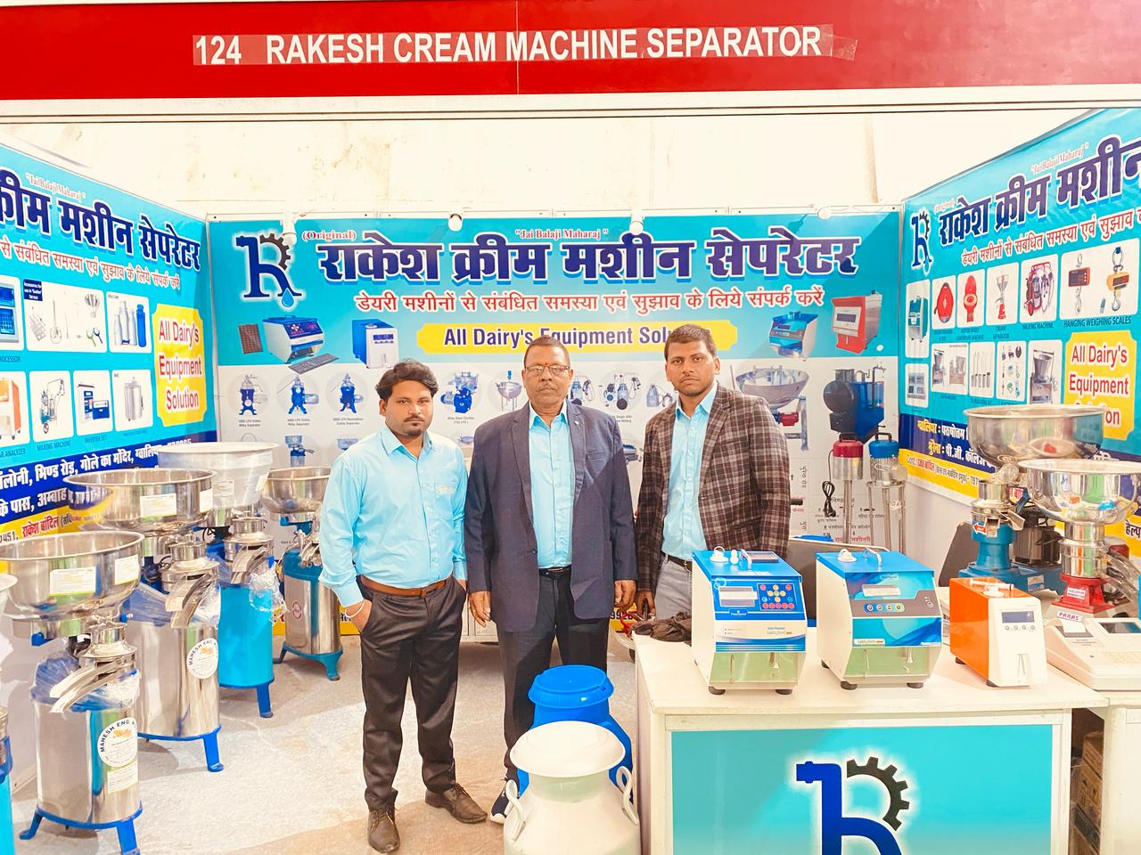 Three men stand proudly in front of a "RAKESH CREAM MACHINE SEPARATOR" booth, displaying dairy equipment.